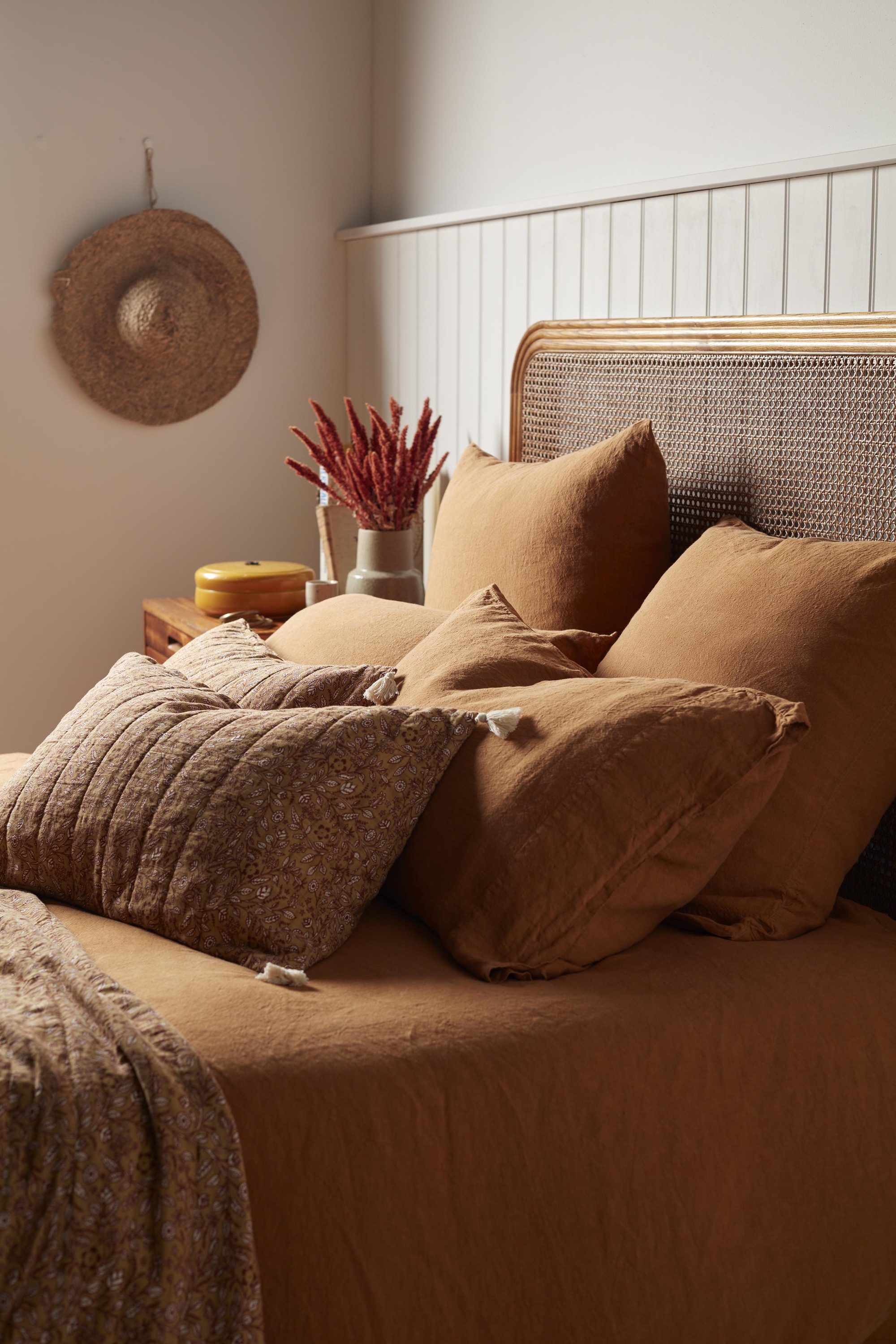 Close-up of a rattan bed with burnt orange bedding. There are four burnt orange pillows and two patterned pillows on the bed.