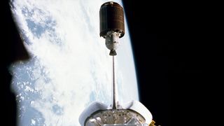 a barrel-shaped satellite is seen departing the payload bay of a space shuttle in Earth orbit
