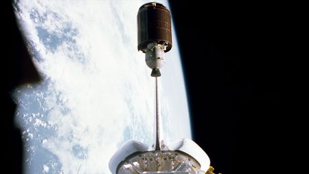 a barrel-shaped satellite is seen departing the payload bay of a space shuttle in Earth orbit
