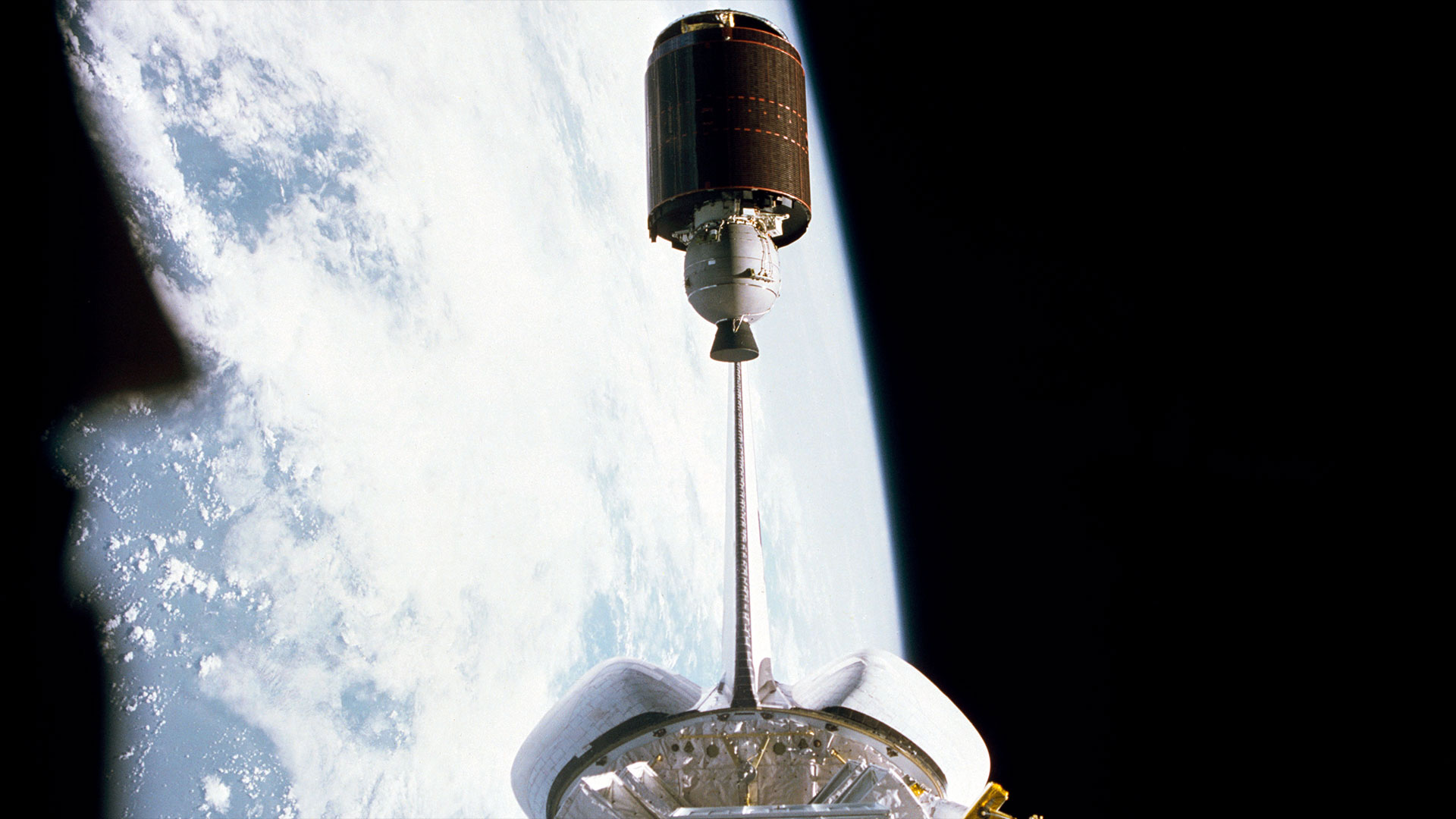a barrel-shaped satellite is seen departing the payload bay of a space shuttle in Earth orbit
