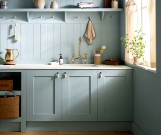 Light blue/green shaker style kitchen cabinets along paneled wall with sink, brass tap and shelf