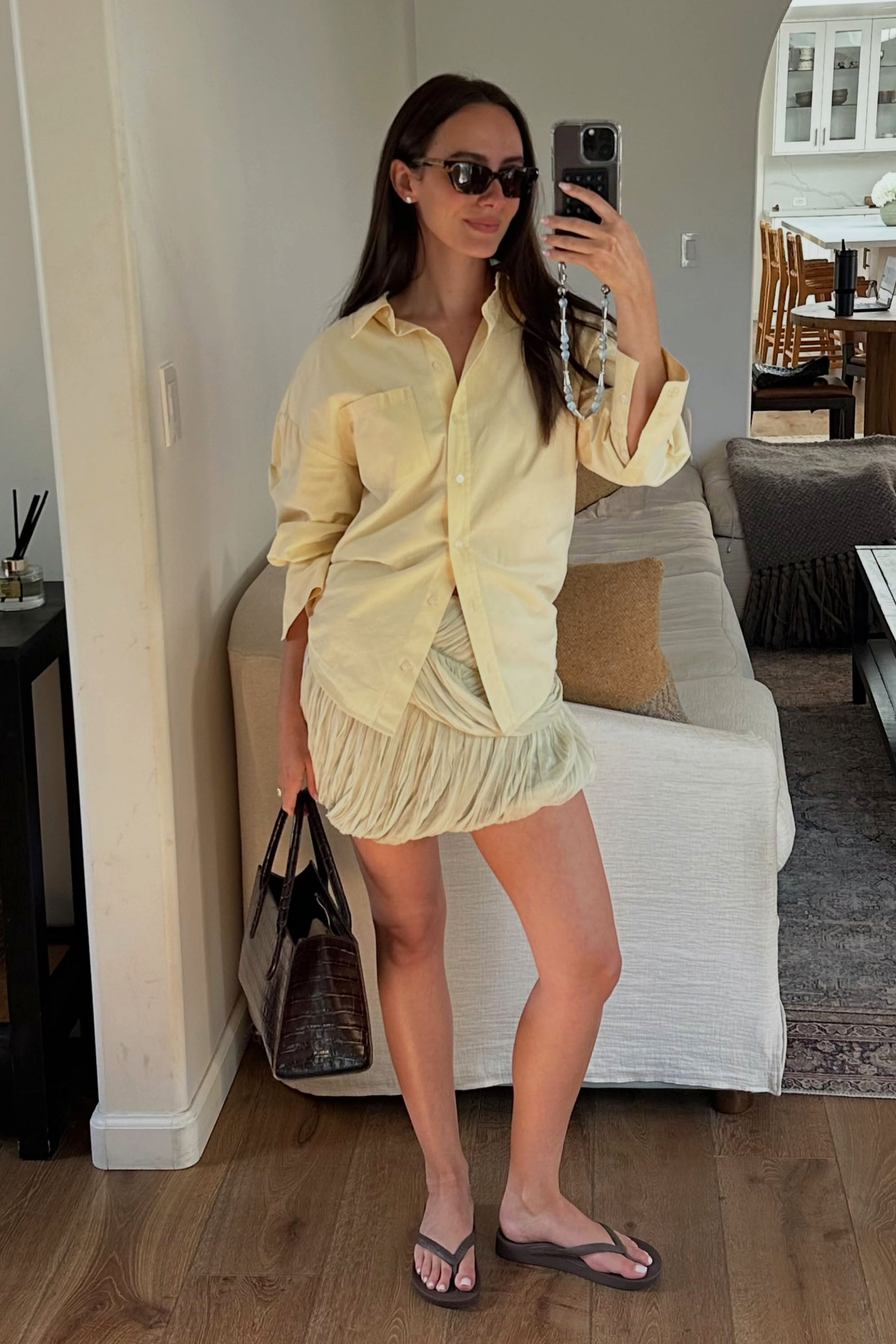 A woman in a butter yellow button-down, skirt, and brown flip flops.