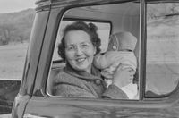 A smiling woman wearing glasses sits in the open window of a car, holding up a bundled infant against a wintry rural backdrop.