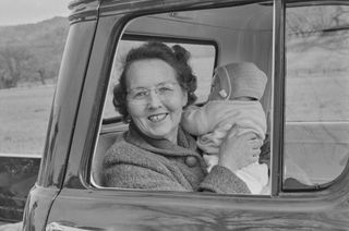 A smiling woman wearing glasses sits in the open window of a car, holding up a bundled infant against a wintry rural backdrop.
