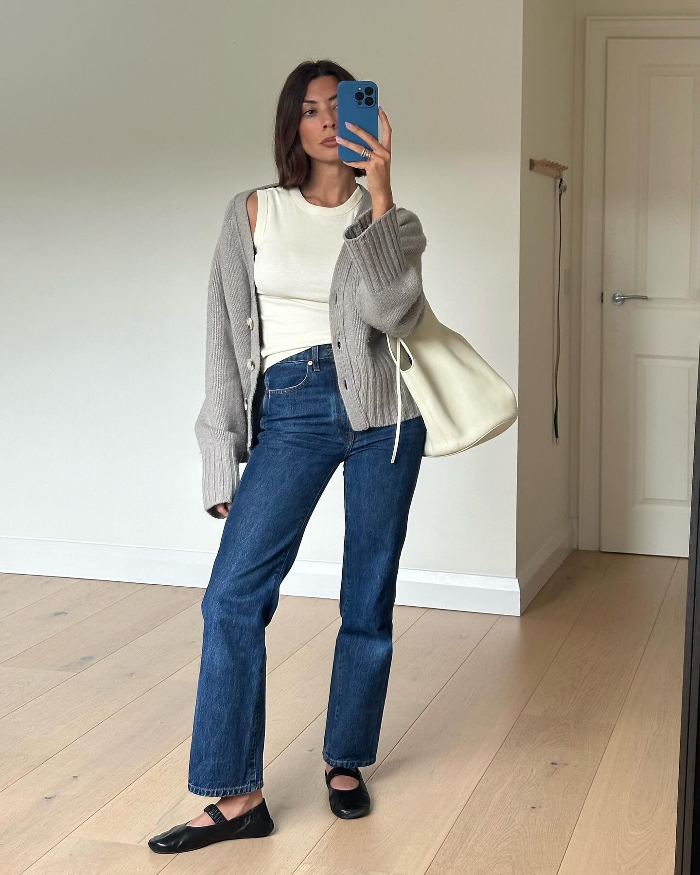 fashion influencer Marianne Smyth poses for a mirror selfie with a blue iPhone case, gray cardigan, ivory tank top, cream leather tote bag, dark wash jeans, and black Mary-Jane flats