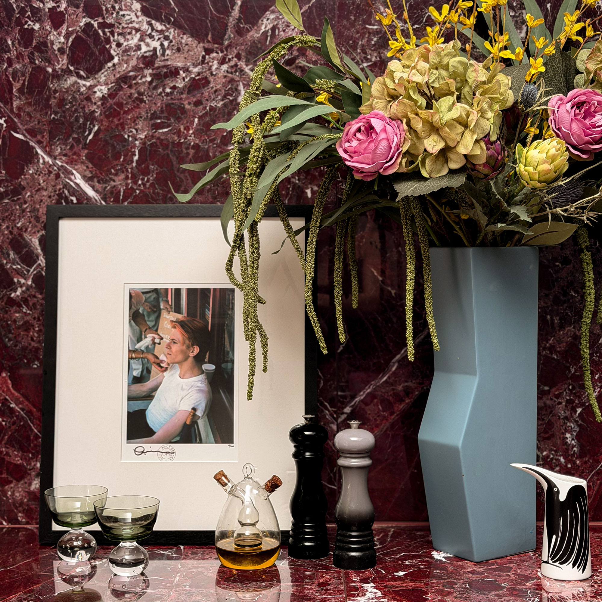 Framed photo, two small glasses, glass oil and vinegar jar, grey salt and pepper grinders and abstract blue vase full of flowers all sitting on red marble