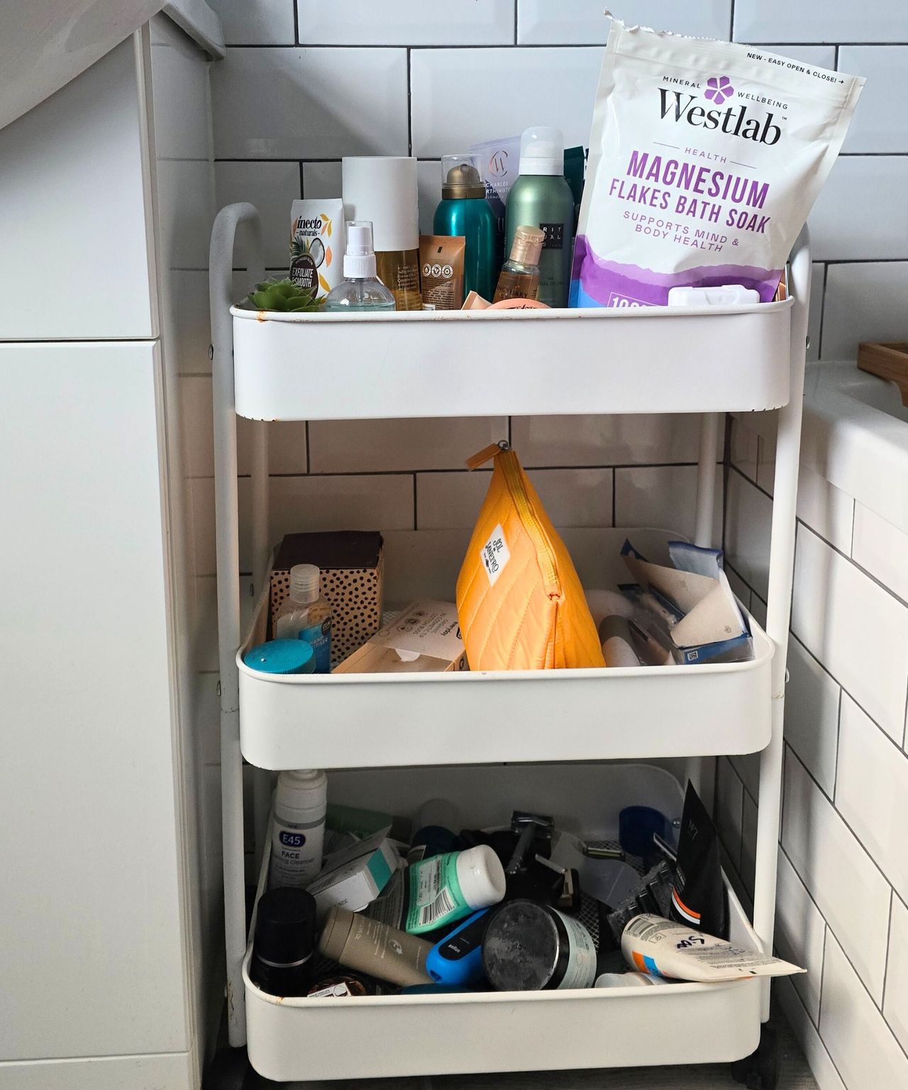 A cluttered three-tier white storage trolley wih colorful toiletries piled on it and white tiles behind it
