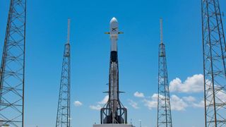 A black and white rocket topped by a payload fairing stands on the launch pad set against a blue sky with a few white clouds