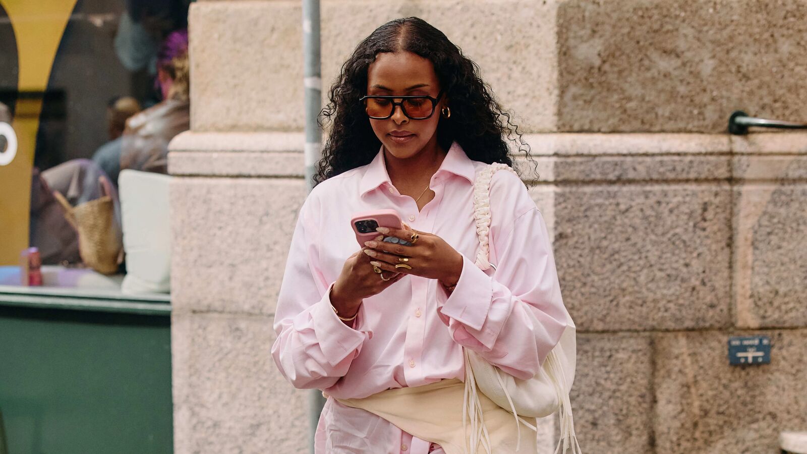 A woman in a pink button-down, white shoulder bag, sweater tied around the waist, and sunglasses, looking at their phone