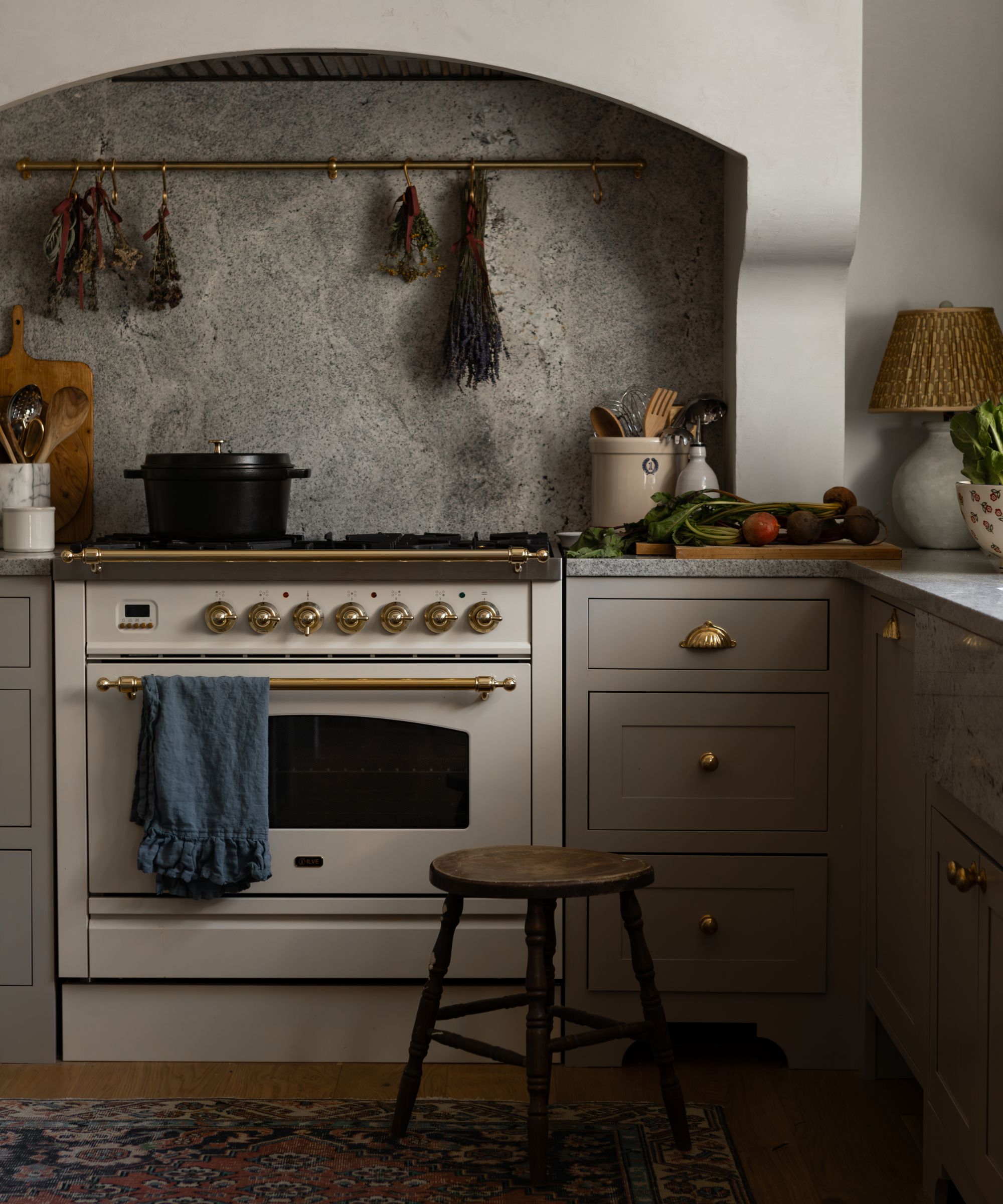 Transitional kitchen with taupe cabinets, a cream cooker, a stone backsplash with an arched top, and brass fixtures. Rustic decor on display, including a woven lamp, dried flowers, and a wood stool.