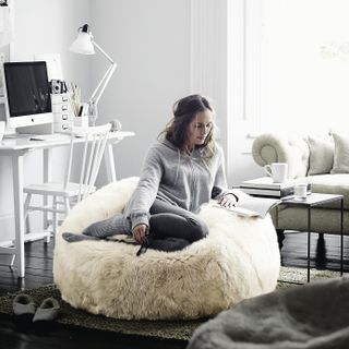 A woman sits reading on a white long haired bean bag in a white living room with dark wooden floors