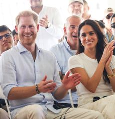 Prince Harry and Meghan Markle smiling and clapping while sitting in chairs