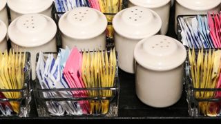 yellow, pink and blue packets of artificial sweeteners are arranged in small baskets, as used in restaurants, and sitting near salt shakers
