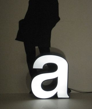 A silhouette of a woman standing behind a large lit up "a" on the ground in front of her.