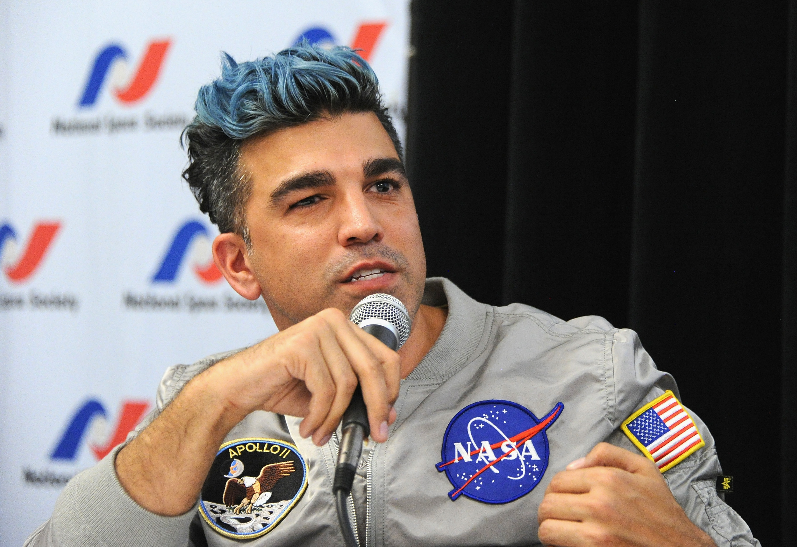 a man with a mohawk in a nasa t-shirt speaks into a microphone while seated at a table on a stage