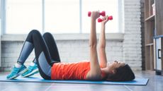 woman on the floor on an exercise mat, knees bent holding a light red dumbbell in each hand, raised overhead over her chest. there's a white brick wall and window behind her. 