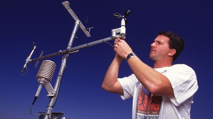 A man wearing a white t-shirt and watch finishes assembling the metal arm on a weather device, made of various metal arms