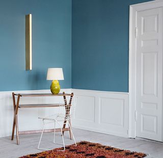 View of a 2013 space at The Apartment gallery featuring blue walls with white wood panelling at the bottom, a brown desk with a green and white lamp on top, a wall light, a white grid style chair, white doors and a multicoloured rug