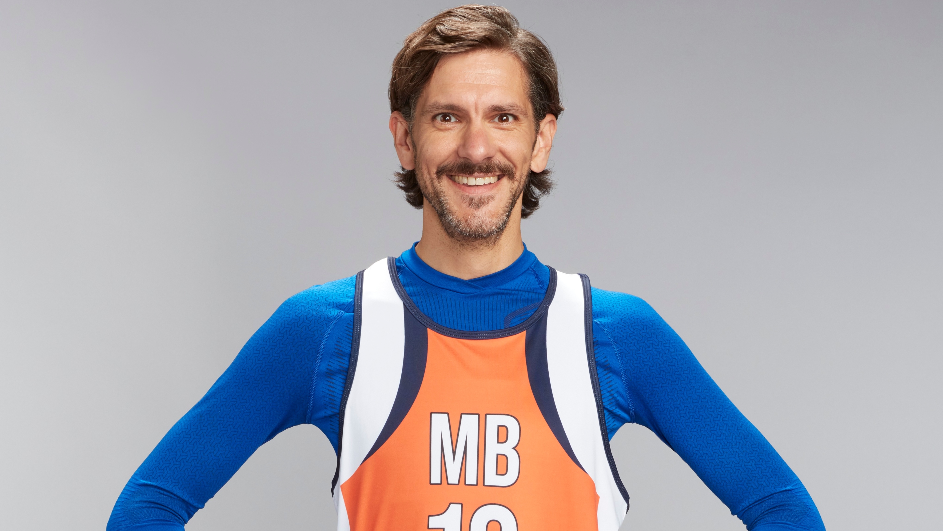 A picture of Mathew Baynton standing against a grey backdrop. Due to the way the image has been cropped, he is visible from the mid-torso up only, and he is wearing a deep blue long-sleeved atheltic top, with a runner's vest over it in orange, black and white with "MB 19" on the front.