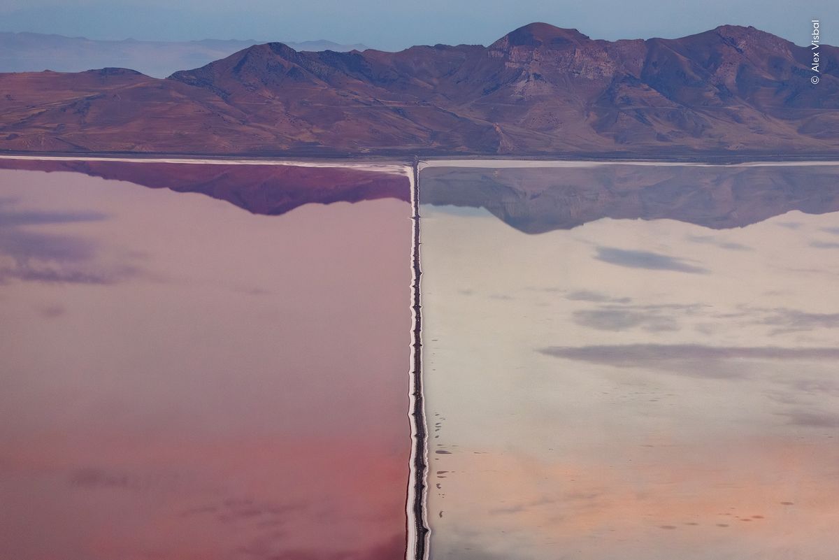 A single engineered line reshaping an entire wetland, waters perfectly still reflecting the mountainous backdrop