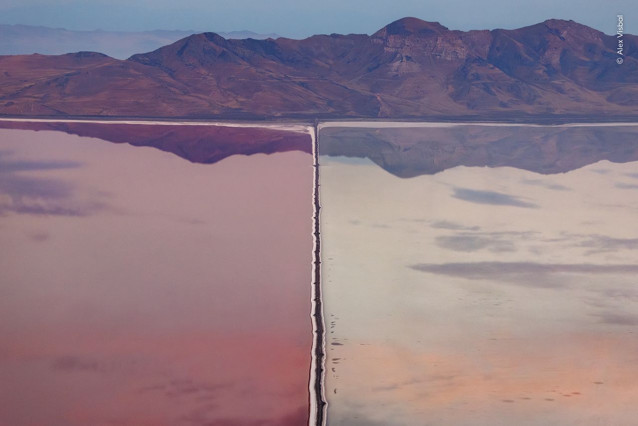 A single engineered line reshaping an entire wetland, waters perfectly still reflecting the mountainous backdrop