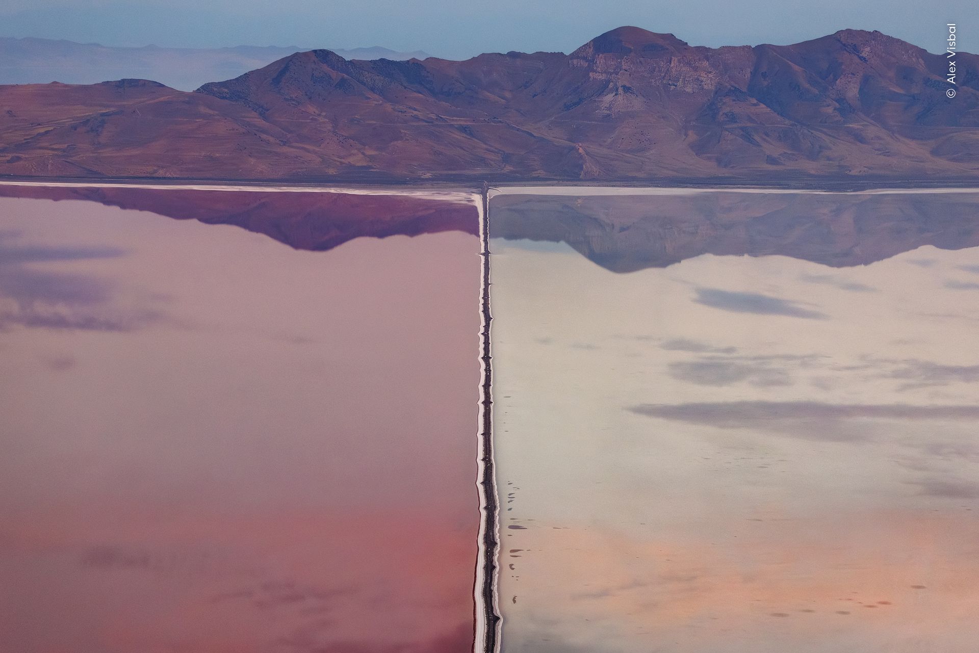 A single engineered line reshaping an entire wetland, waters perfectly still reflecting the mountainous backdrop