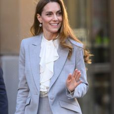 Kate Middlton steps outside wearing a light grey suit with black pointed-toe heels and a cream ruffle blouse.