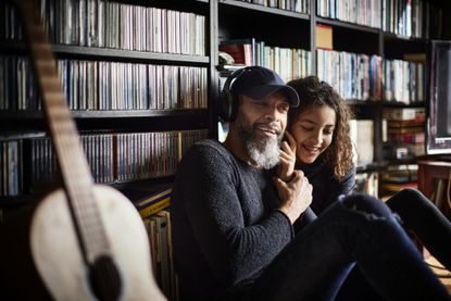 A grandfather listens to music with his granddaughter. They are in a room lined with CDs and books, and guitar is in the foreground.