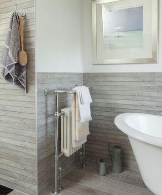 A bathroom with light gray wooden shiplap paneling across half the walls, a gray towel and brush hung up on the wall, a silver towel rail with two white towels hung up on it, and white wall with a framed sea print