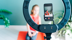 A close up of a ring-shaped light with a phone in the middle filming a girl on a yoga mat behind it.