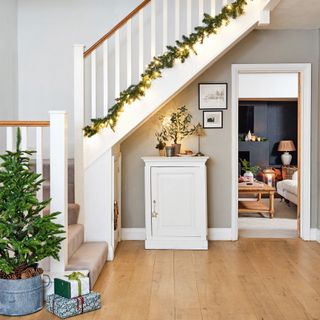 a hallway with a stairwell covered in a fairy lit green garland, with a view through to a cost living room