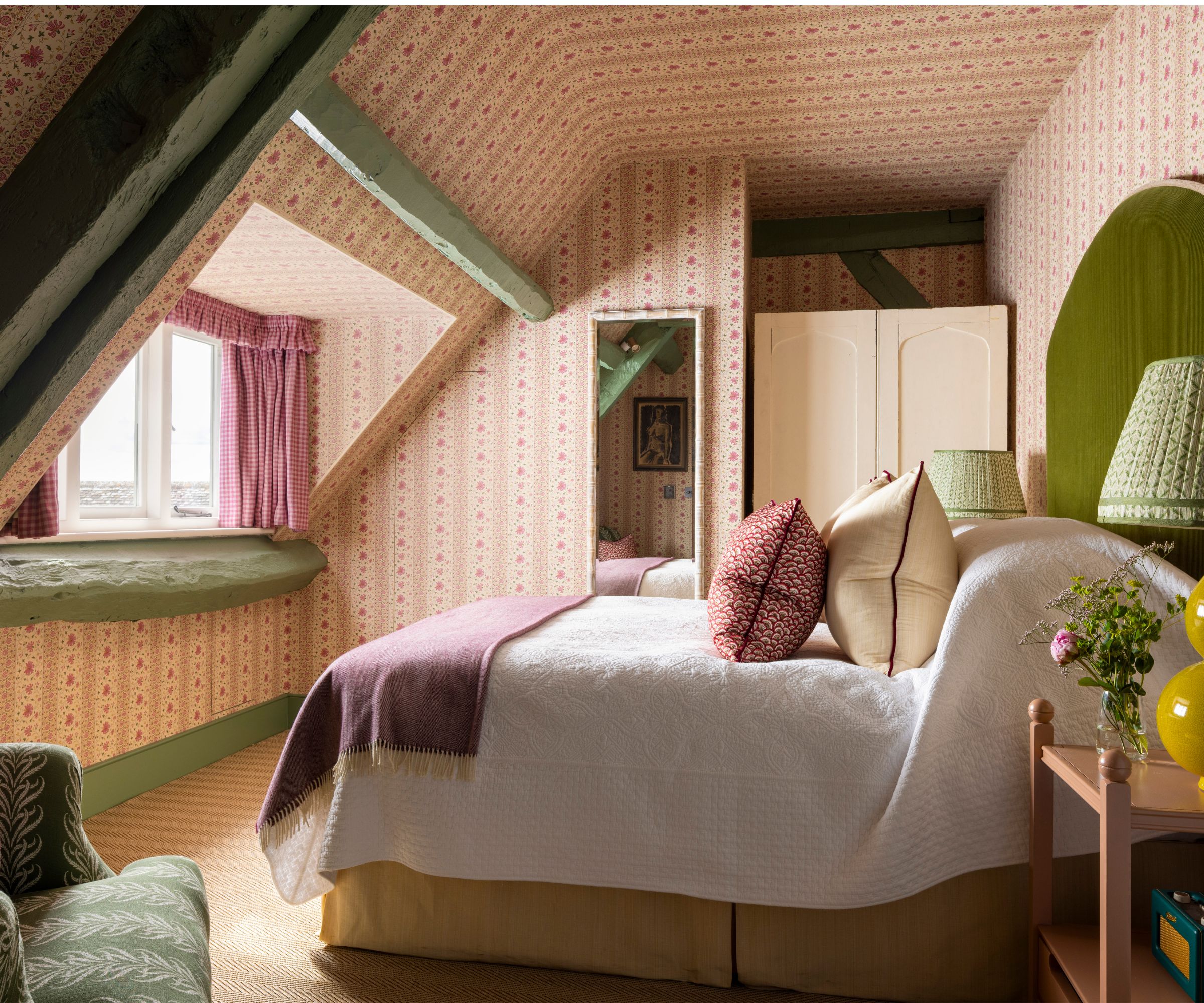 soft pink floral wallpapered attic bedroom with a green velvet headboard, dressed with a large white quilt, yellow bed skirt, pink throw and throw pillows