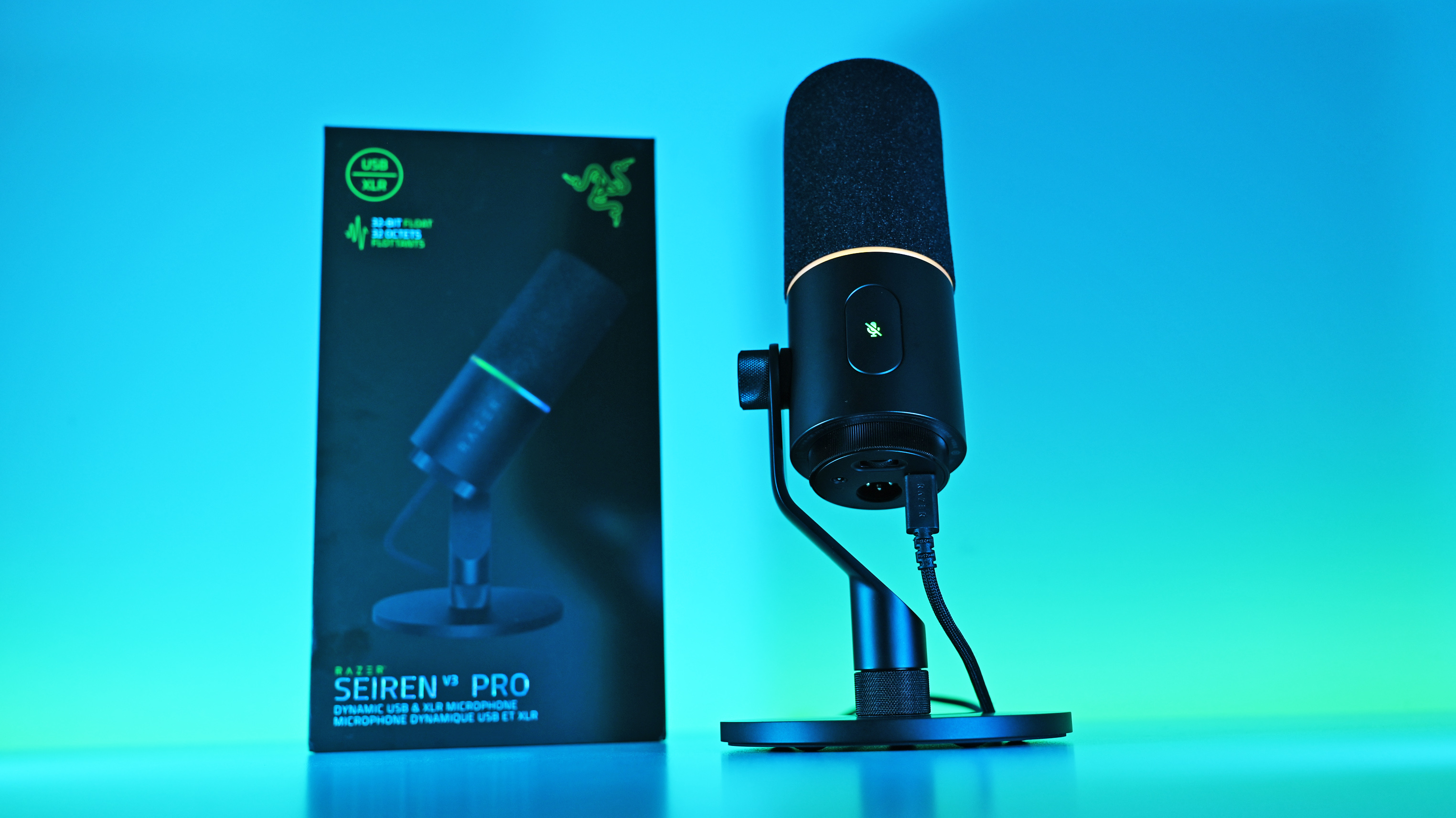 Low-angle shot of the Razer Seiren V3 Pro mic plugged into a USB-C cable next to its box, illuminated by a vivid blue and cyan background.