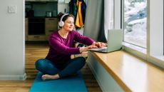 Woman setting up laptop to do low-impact cardio workout at home on yoga mat with snow outside window and wearing headphones