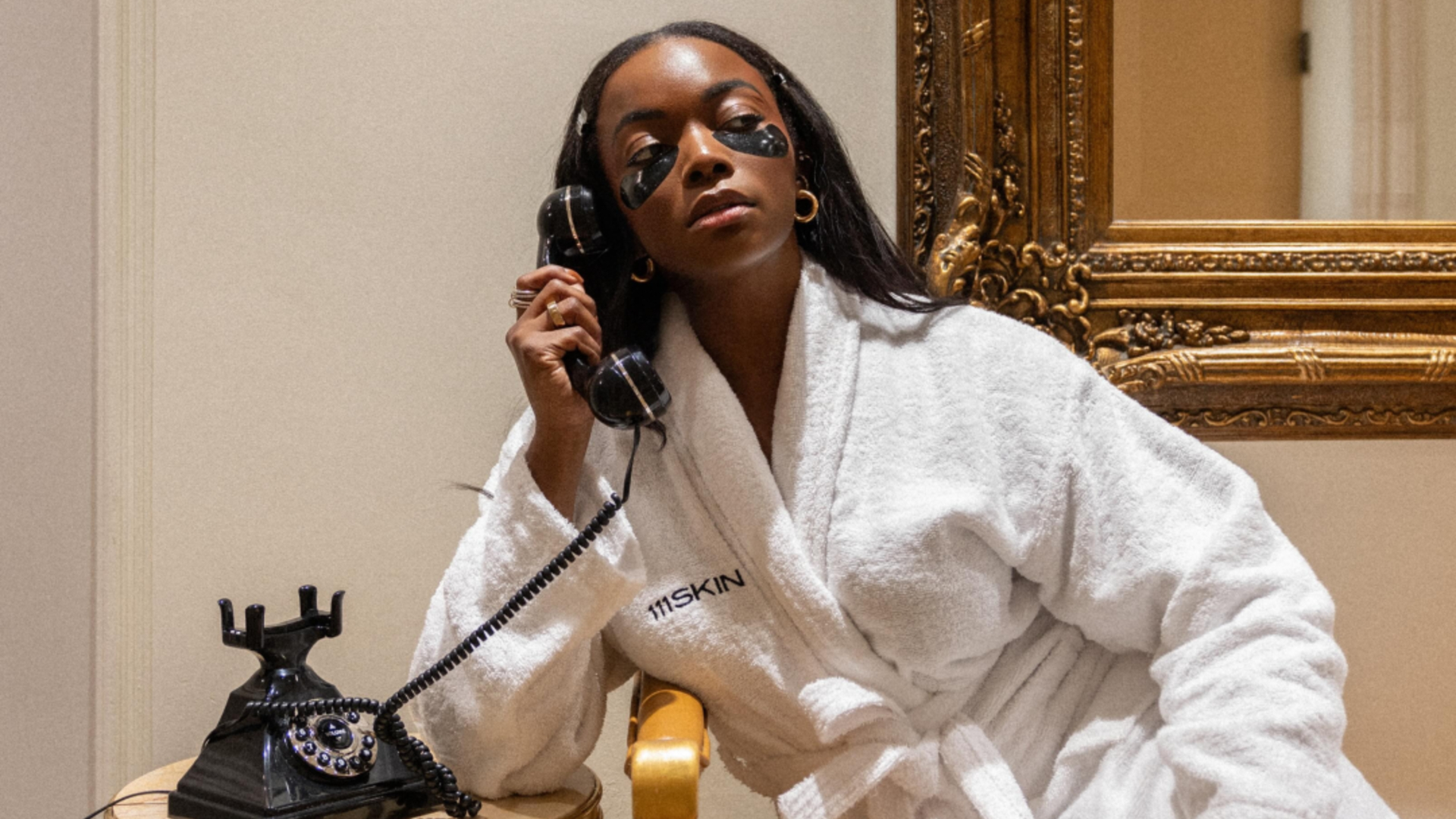 A woman wears skincare eye patches and talks on the phone.