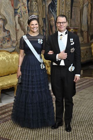 Crown Princess Victoria wearing a navy ballgown standing on the arm of Prince Daniel wearing a black tuxedo and medals in front of a gold sofa