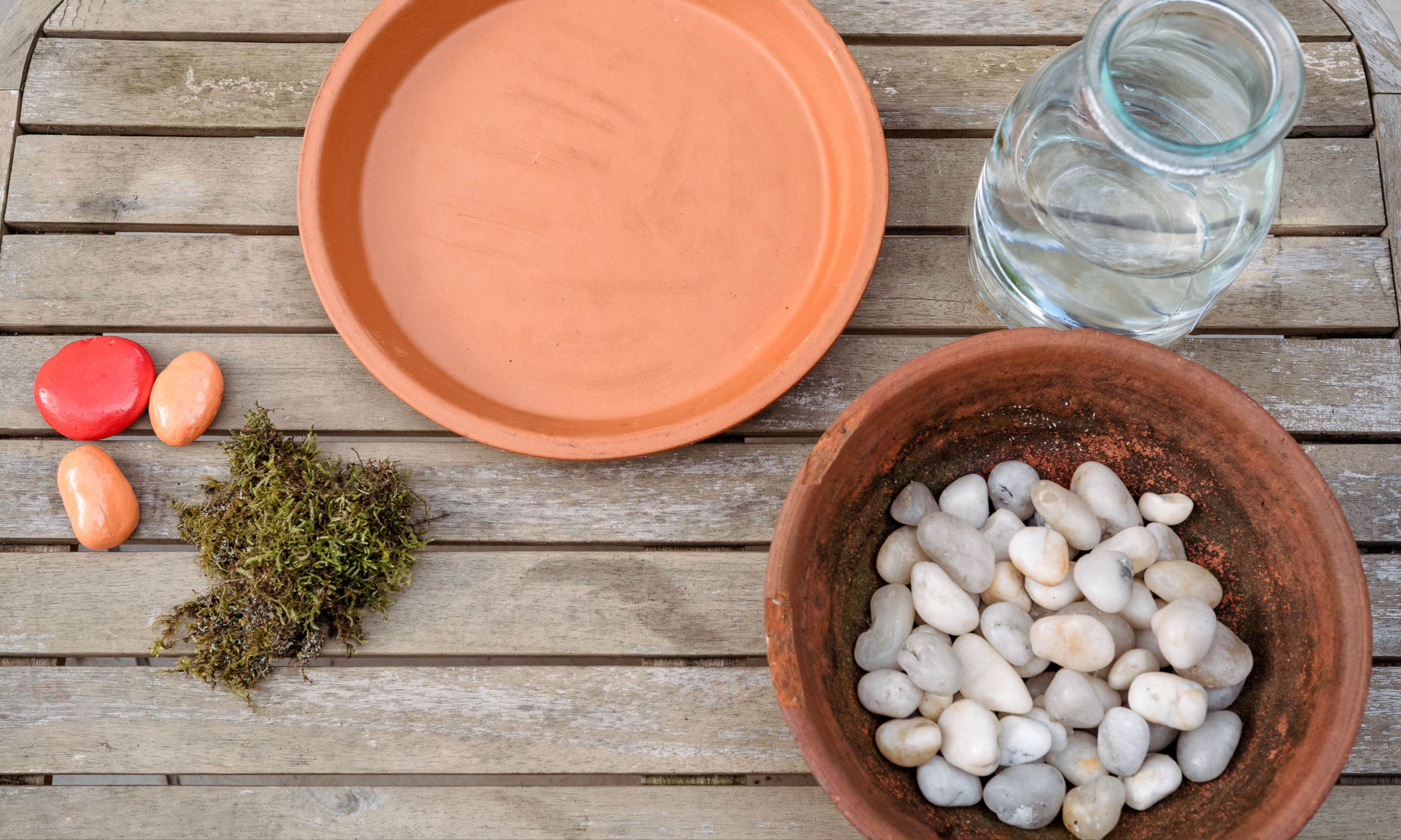 flay lay of glass vase, white stones in rustic bow, terracotta saucer, moss and painted stones