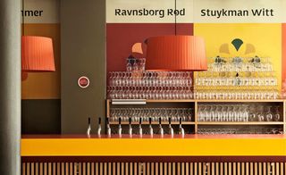 A yellow and red bar counter with a line of beer taps on it, shelves behind it with many glasses on them and a large round orange pendant light hanging above.