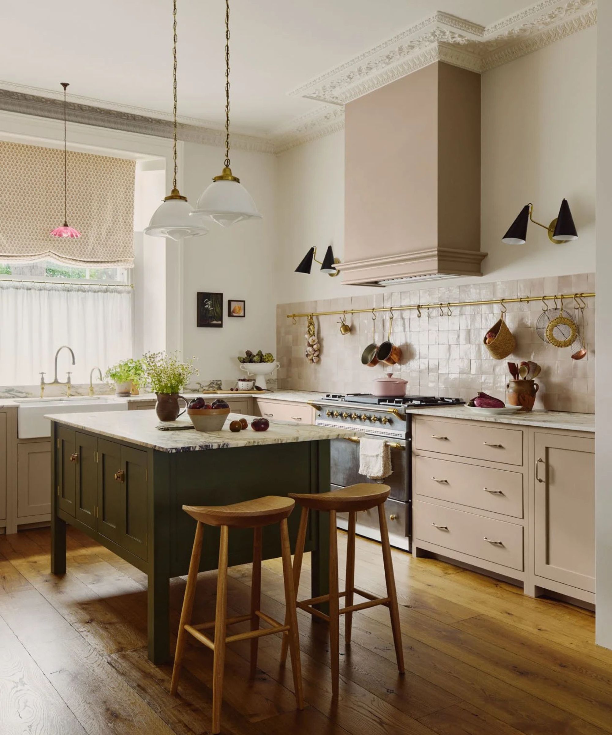 A traditional kitchen with pink cabinets, a green freestanding island, pink backsplash tiles, and a brass pot rack on the wall