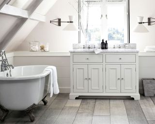 Bright bathroom with gray wooden flooring, white bathroom cabinet with marble countertop and twin sinks, large white freestanding bath, twin white and metal wall lamps over sink space, white painted paneling on walls, integrated wall shelf around the bathroom, decorated with glass accessories