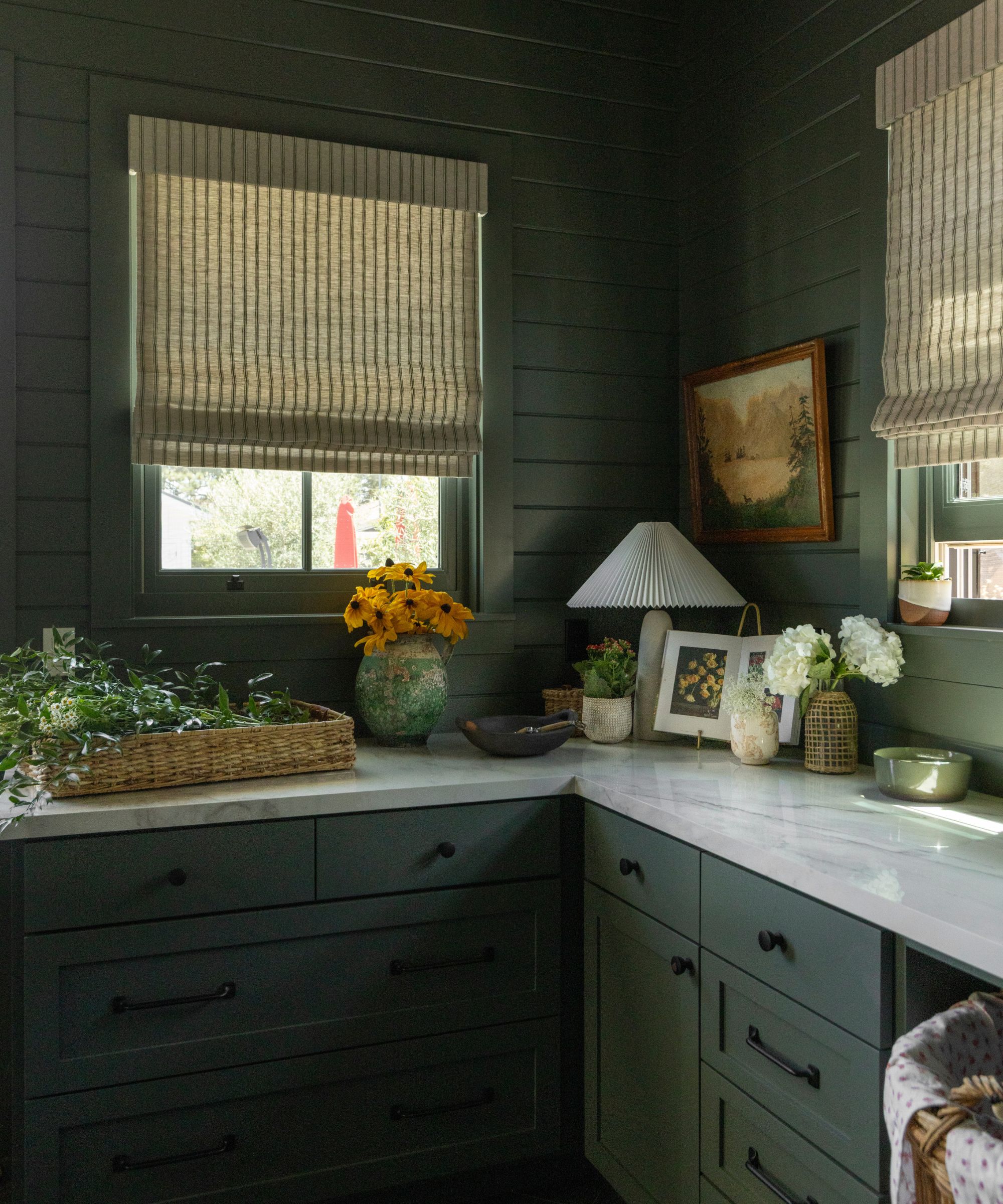 A kitchen with green cabinets, white marble countertops, green panelled walls, and two windows with neutral blinds.