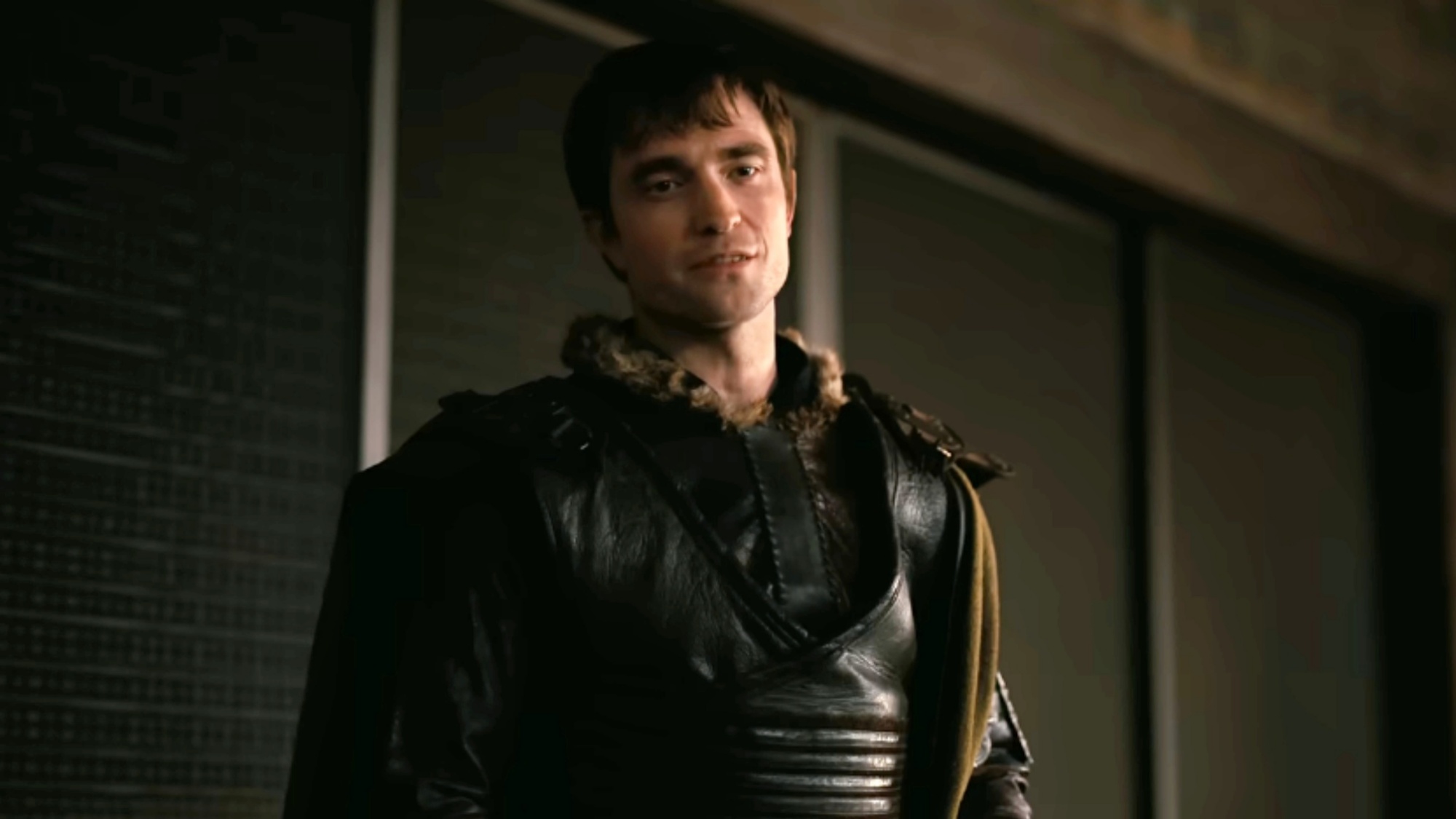 A screen shot of Robert Pattinson as Antinous looking serious while wearing black leather.