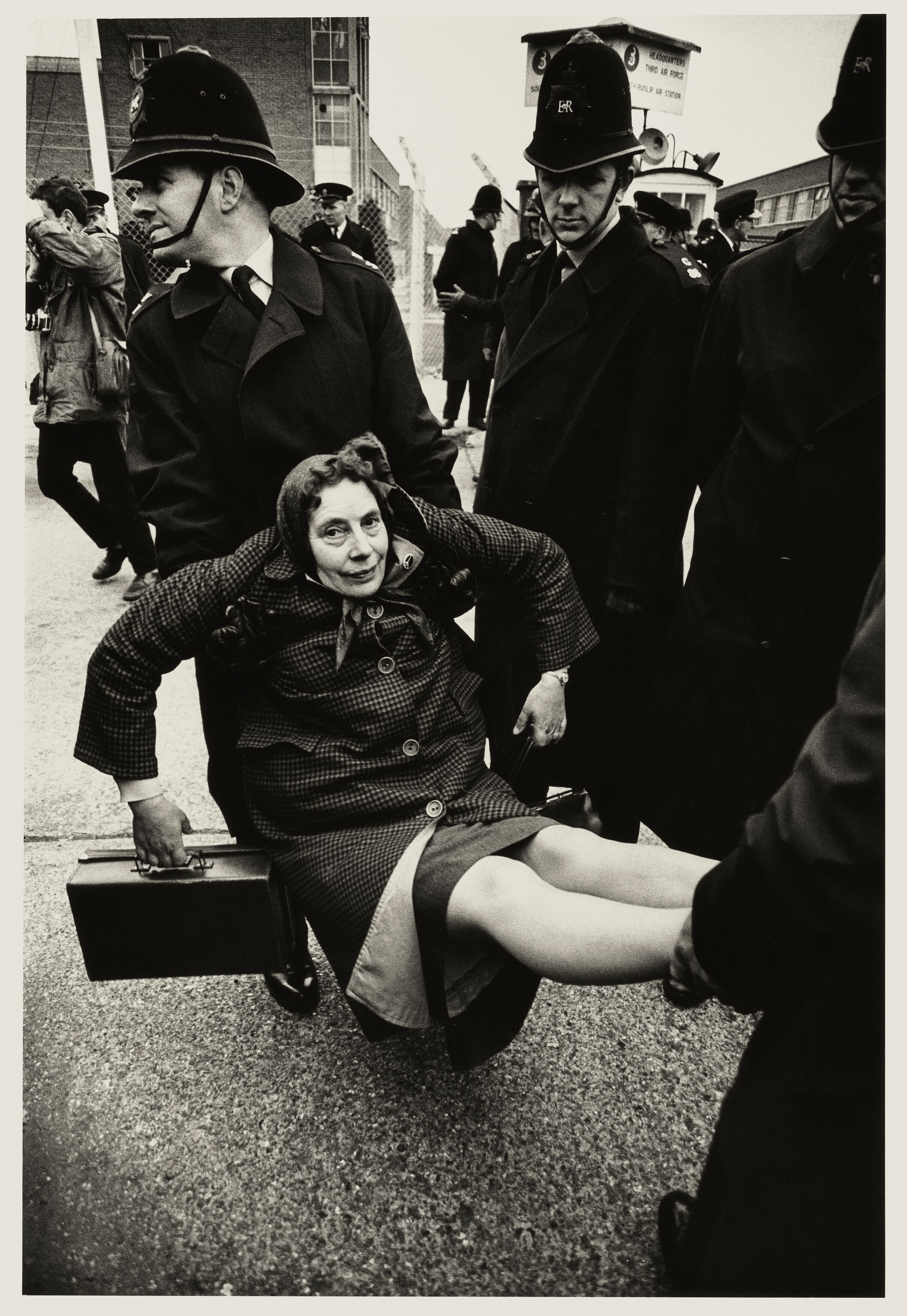 Two British police officers in traditional custodian helmets carry a seated woman away from a protest site near an air station.