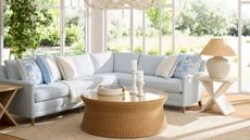 Blue L-shaped sofa with blue and white cushions behind a circular rattan coffee table: in the background, floor-to-ceiling windows looking onto a garden view.