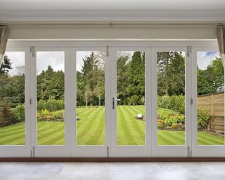 A set of concertina patio doors with silk curtains on either side and a view of a fabulous garden. A large nicely mowed lawn stretches away towards a backdrop of mixed trees