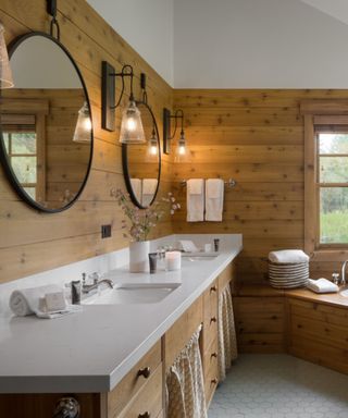A light wooden bathroom area with two circular black mirrors, two glass wall sconces, and a white sink unit with towels and candles on it with light wooden drawers and a corner seat with towels on to the right