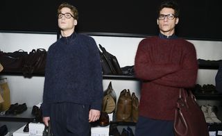 Two male models wearing looks from Giorgio Armani's collection. Both models are wearing blue trousers with one model wearing a blue jumper and another model wearing a wine coloured jumper and holding a similar coloured bag. They are both wearing glasses and behind them are shelves with multiple bags and shoes