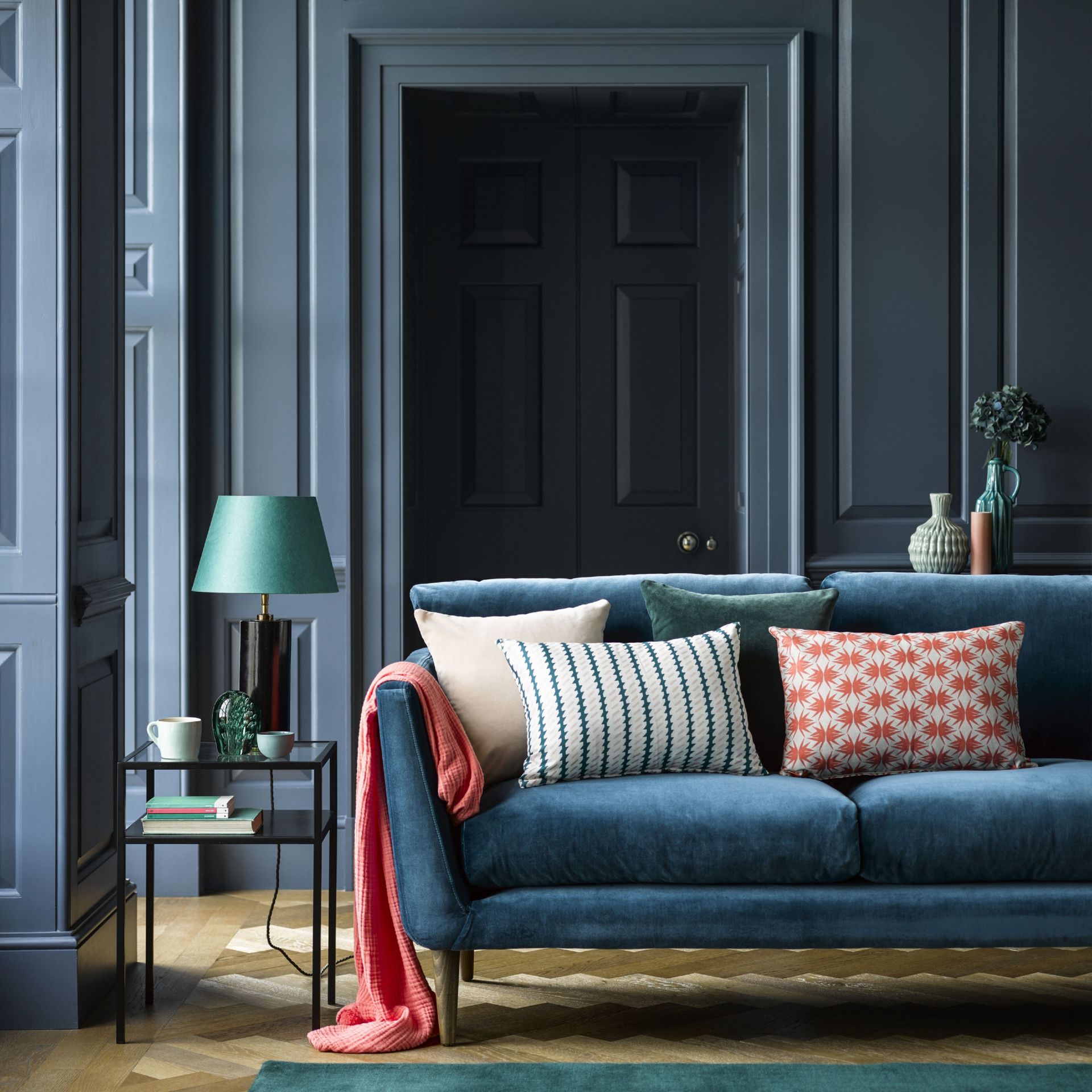 Skirting color ideas, living room painted blue with matching paneling, woodwork and walls, blue sofa, hardwood floor, black and metal end table, coral blanket and pillows, blue/emerald pillows and rug