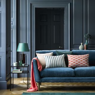 skirting board colour ideas, blue painted living room with panelling, matching woodwork and walls, blue sofa, hardwood floor, black and metal side table, coral throw and cushions, blue/emerald cushions and rug
