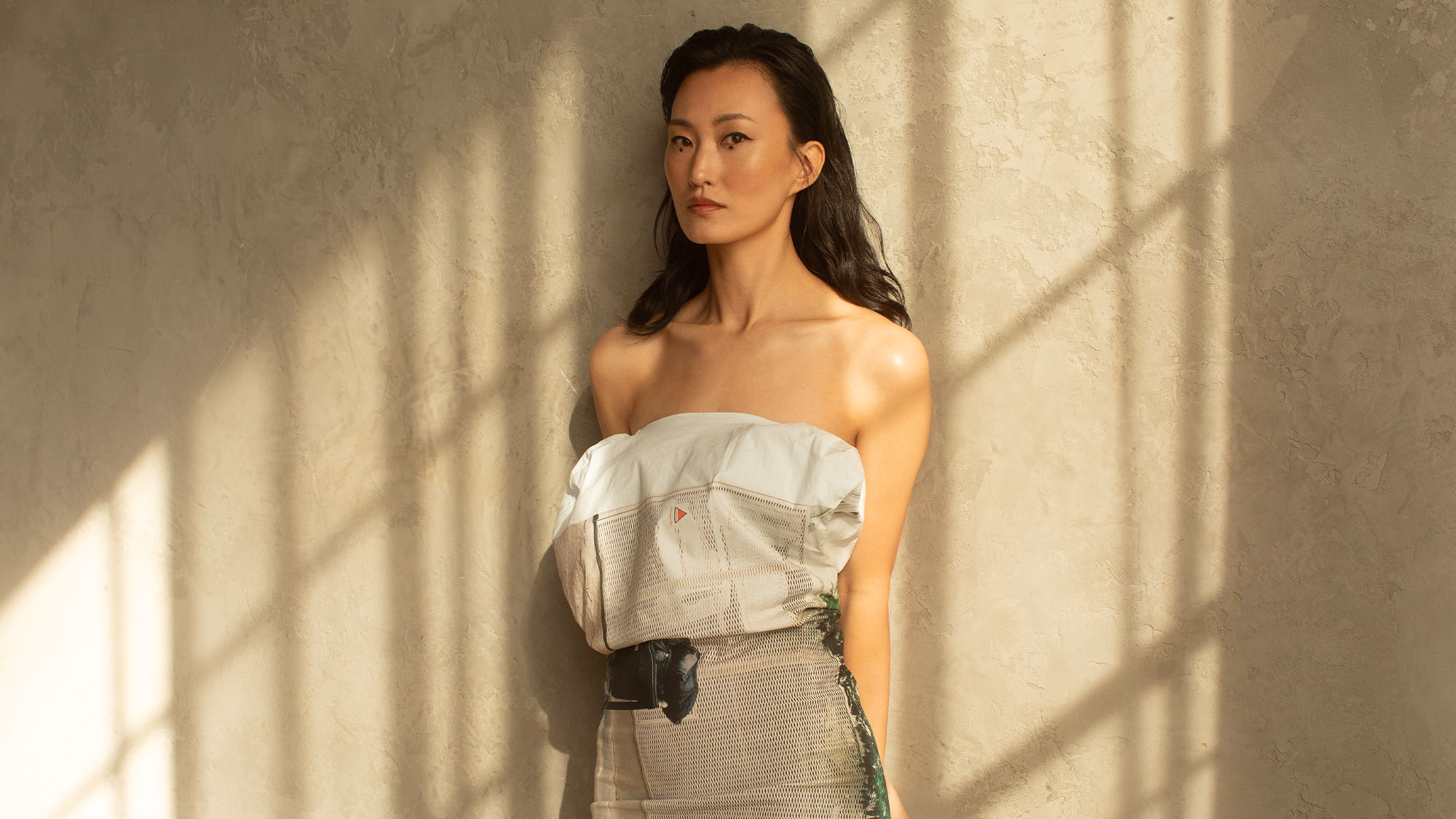 Actress Mari Yamamoto standing in front of a textured white wall. She is wearing a printed strapless dress. 